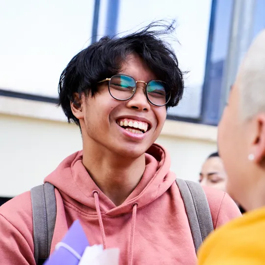 A young man in a pink hoodie and glasses laughs while talking with a classmate outdoors, with his dark hair blowing in the wind.
