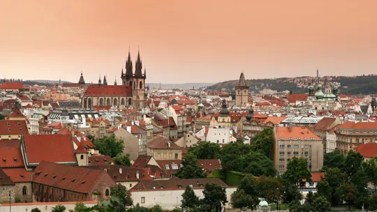 Prague cityscape with red rooftops and gothic church spires under a peach-coloured sky
