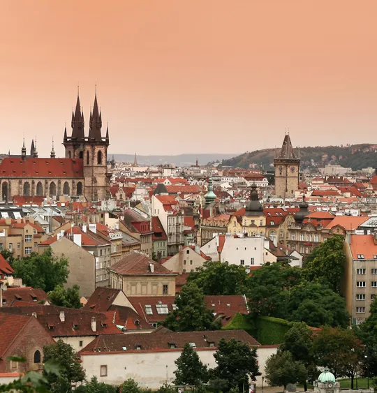 Prague cityscape with red rooftops and gothic church spires under a peach-coloured sky
