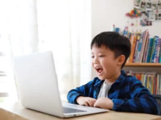 A photograph of a very young boy with short black hair smiling at a laptop computer whilst sitting at a table.