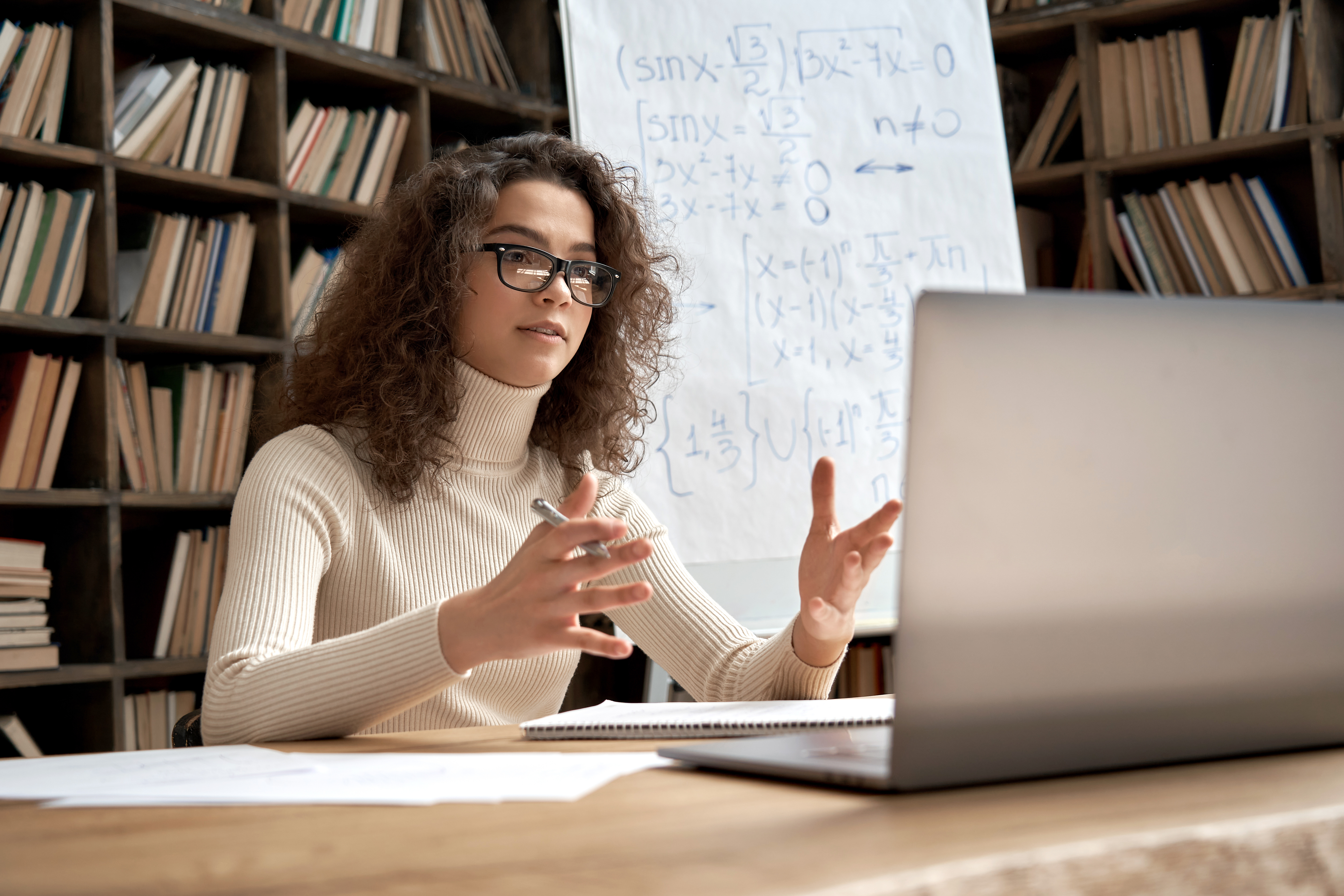 A specialist online tutor gestures towards her laptop as she teaches a complex maths lesson from her study.