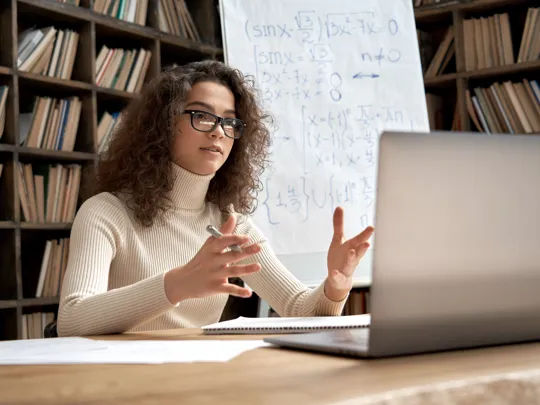 A specialist online tutor gestures towards her laptop as she teaches a complex maths lesson from her study.