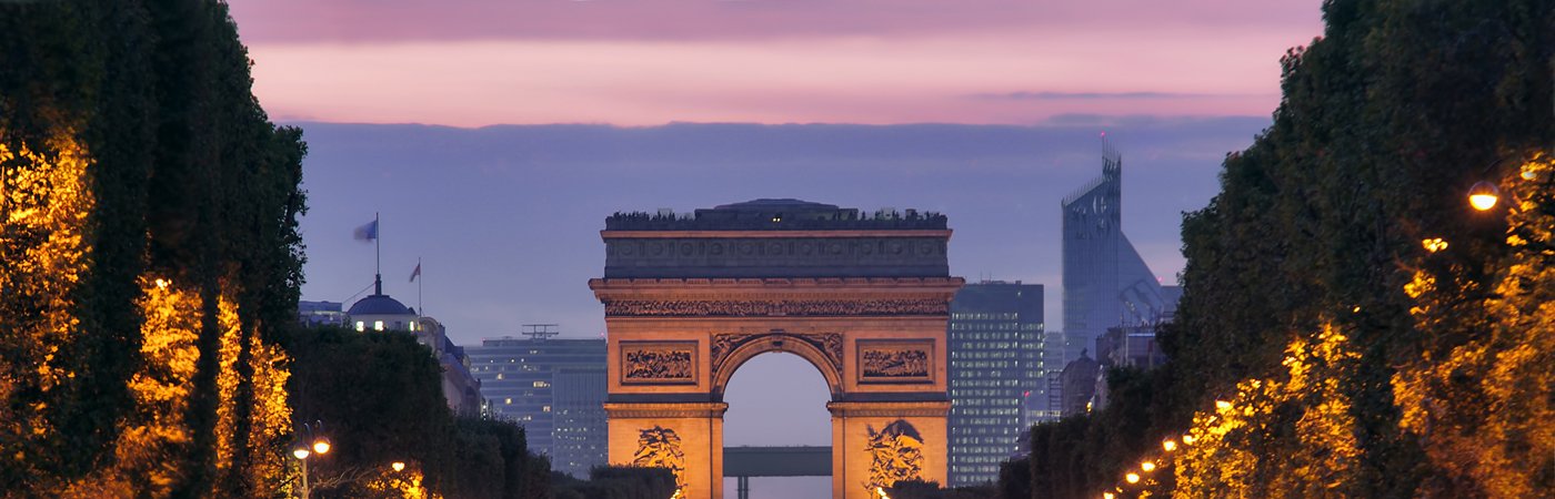 Arc de Triomphe seen from the Champs-Élysées at twilight, with city lights and skyscrapers in the background.