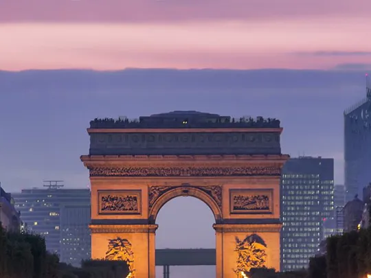 Arc de Triomphe seen from the Champs-Élysées at twilight, with city lights and skyscrapers in the background.