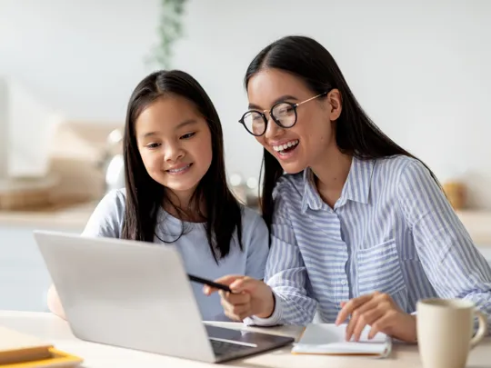 An parent guides a child using a laptop during a homeschooling session in Kuala Lumpur, both smiling and looking at the screen.