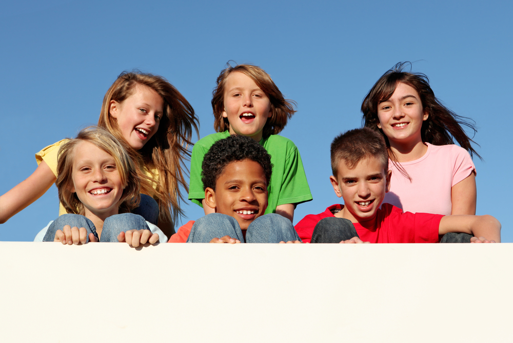 Happy group of children leaning over a wall, smiling under a clear blue sky.