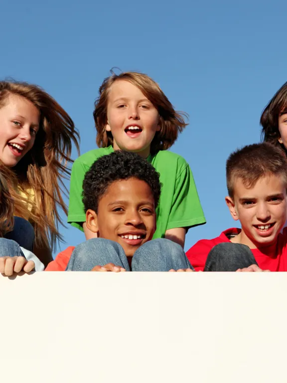 Happy group of children leaning over a wall, smiling under a clear blue sky.