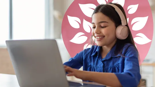 A young student wearing pink headphones, smiling and looking at a laptop while typing.
