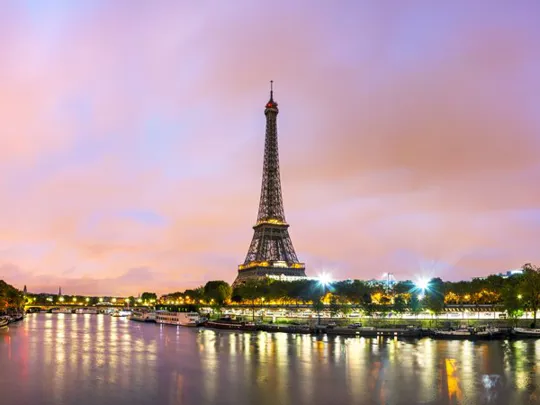 Eiffel Tower illuminated at dusk, reflected in the River Seine under a pastel sky in Paris.