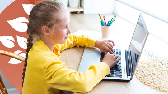 A young student with braided hair, wearing a yellow top, looking at a laptop and typing at a wooden desk.