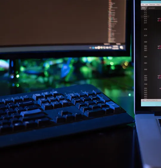 A young woman in a green sweater and glasses smiles while focused on working on a silver laptop in a brightly lit, modern indoor public area.