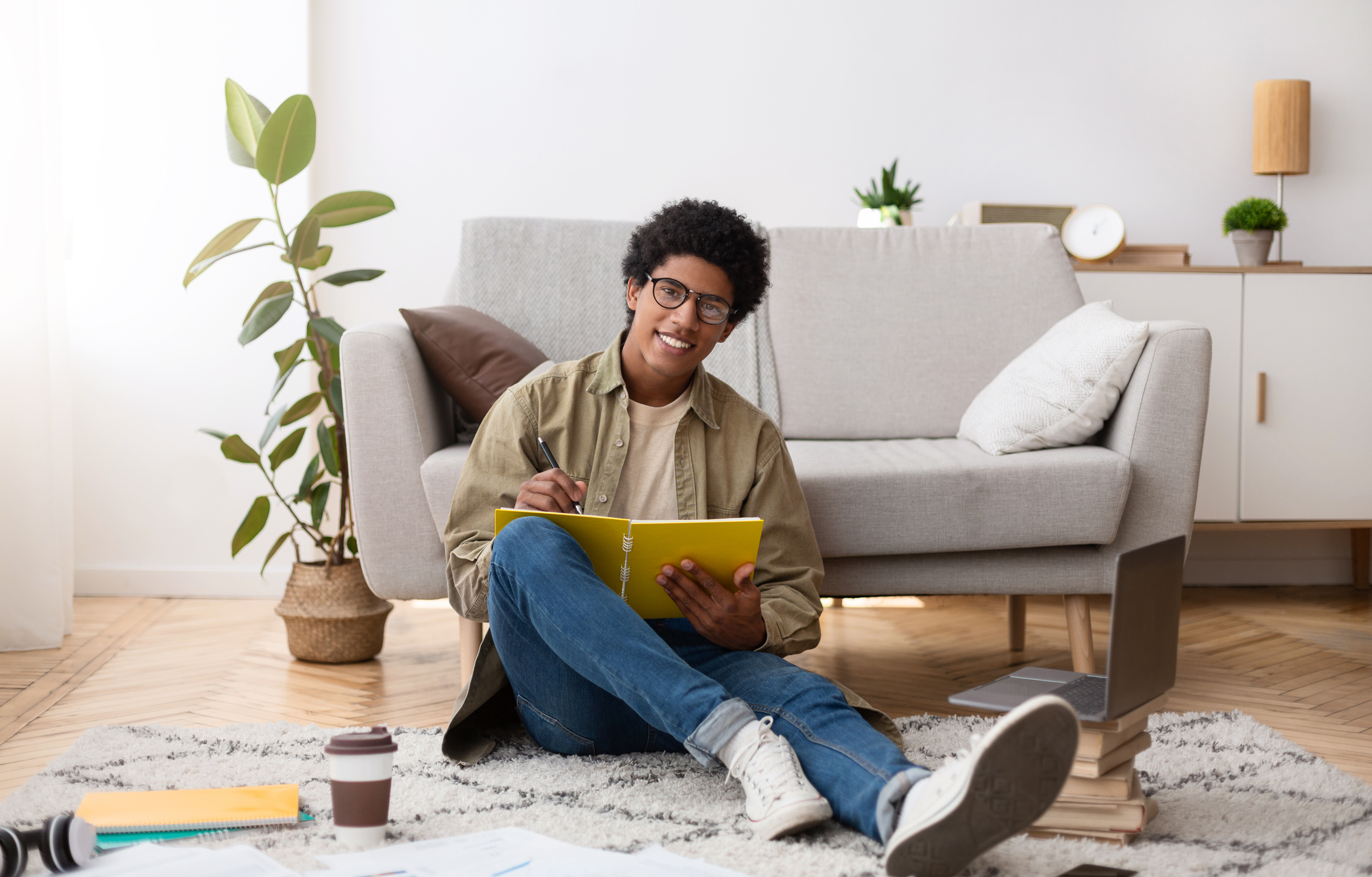 A smiling male student with glasses sits on the floor of a living room, writing in a notebook with a laptop and books nearby, creating a comfortable study environment.