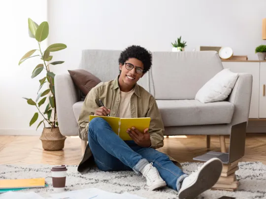 A smiling male student with glasses sits on the floor of a living room, writing in a notebook with a laptop and books nearby, creating a comfortable study environment.