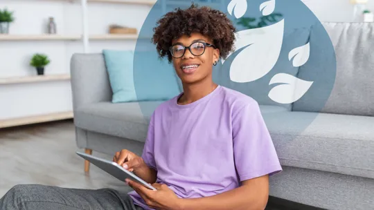 A teenager with glasses and braces, smiling and looking at the camera while holding a tablet, sitting on the floor in a casual setting.