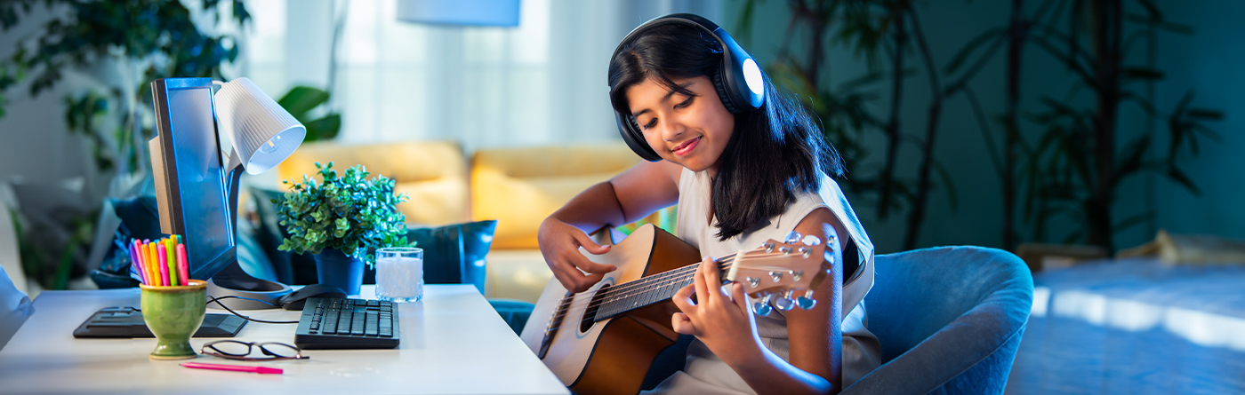 Smiling girl playing acoustic guitar with headphones on, seated at a desk during an online lesson.