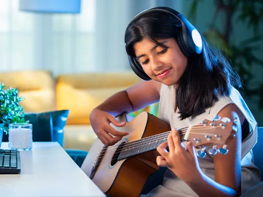 Smiling girl playing acoustic guitar with headphones on, seated at a desk during an online lesson.