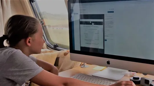 Learner engaging with online coursework on a desktop computer in a well-lit caravan setting.