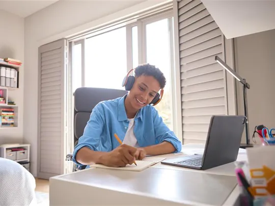 A smiling teenage student wearing headphones studies at a desk with a laptop, studying his GCSE Level courses from home.