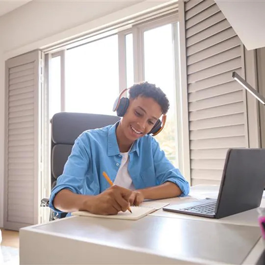A smiling teenage student wearing headphones studies at a desk with a laptop, studying his GCSE Level courses from home.
