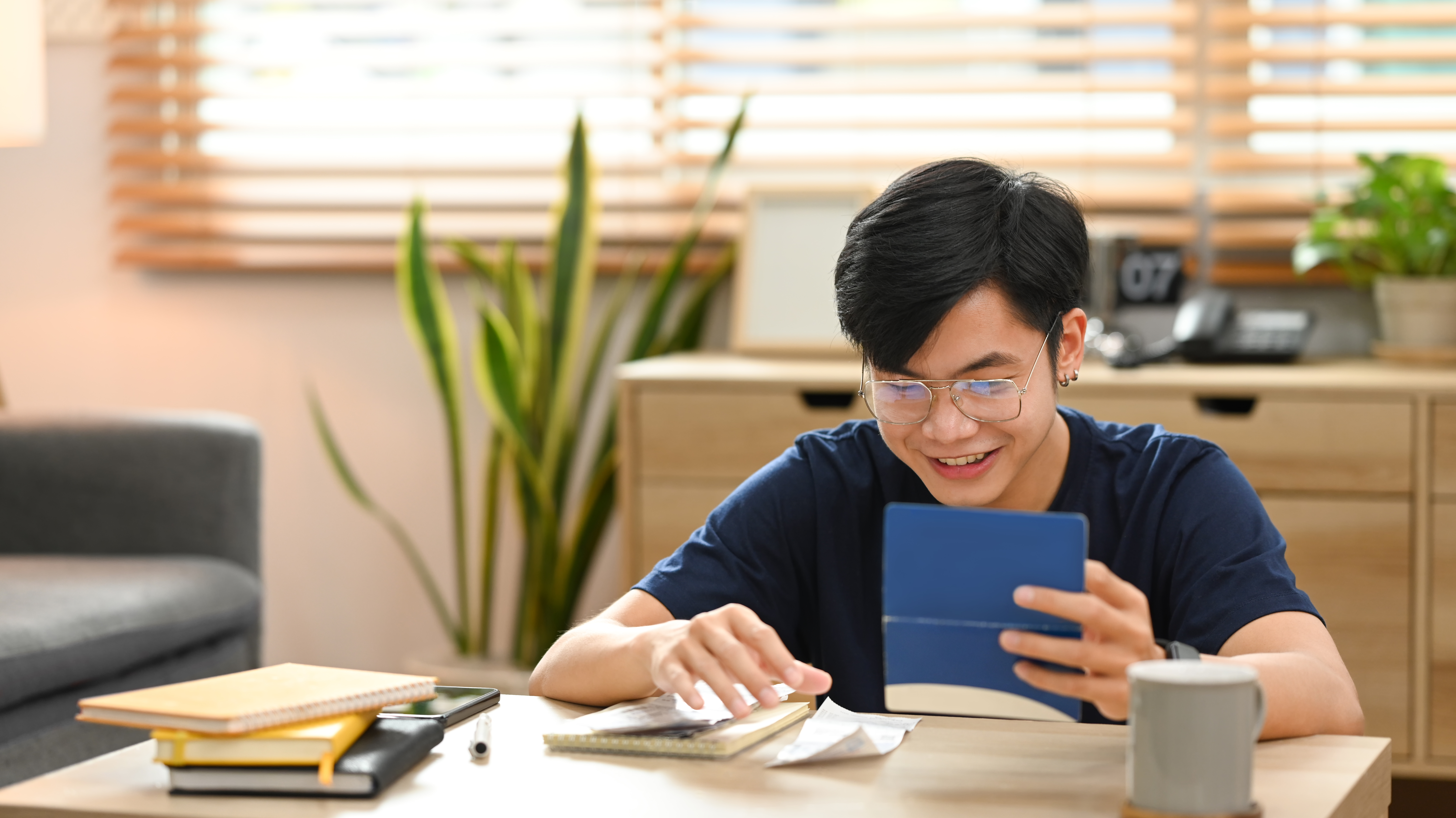 A young male student with glasses smiles while studying at his desk, using a digital tablet and notebooks in a bright, modern room.