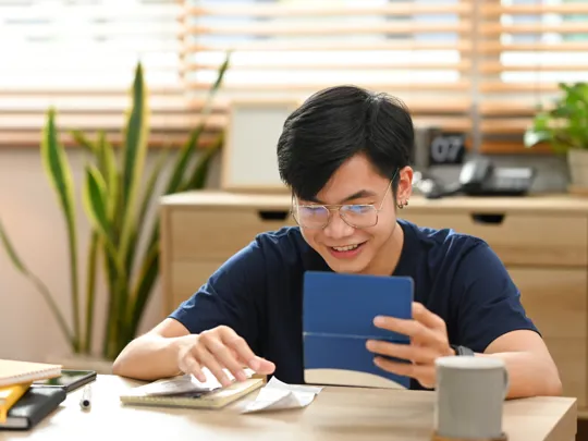 A young male student with glasses smiles while studying at his desk, using a digital tablet and notebooks in a bright, modern room.