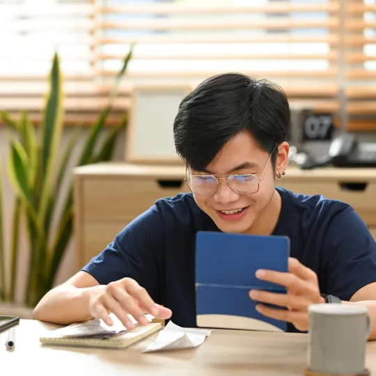 A young male student with glasses smiles while studying at his desk, using a digital tablet and notebooks in a bright, modern room.