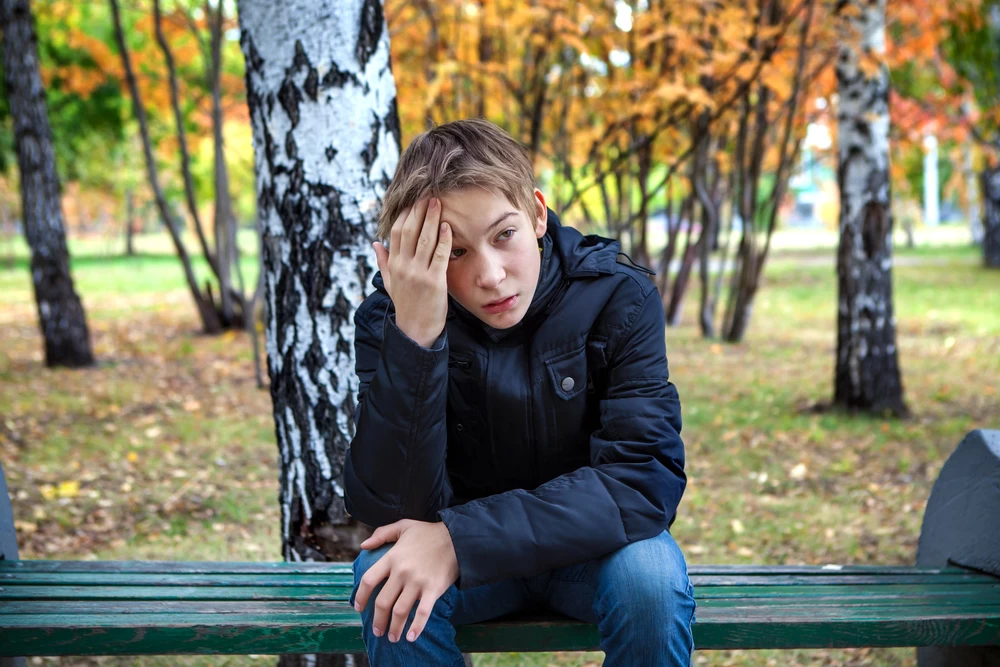 Young boy sat outside on a bench with his hand on his head. 