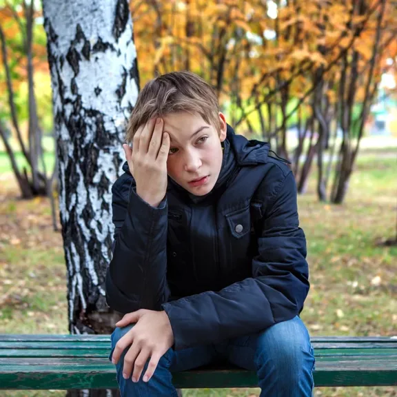 Young boy sat outside on a bench with his hand on his head.