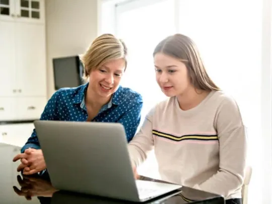 A homeschooling mother and a daughter looking at a laptop at their kitchen table.