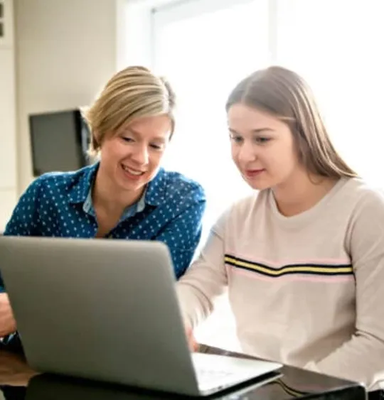 A homeschooling mother and a daughter looking at a laptop at their kitchen table.