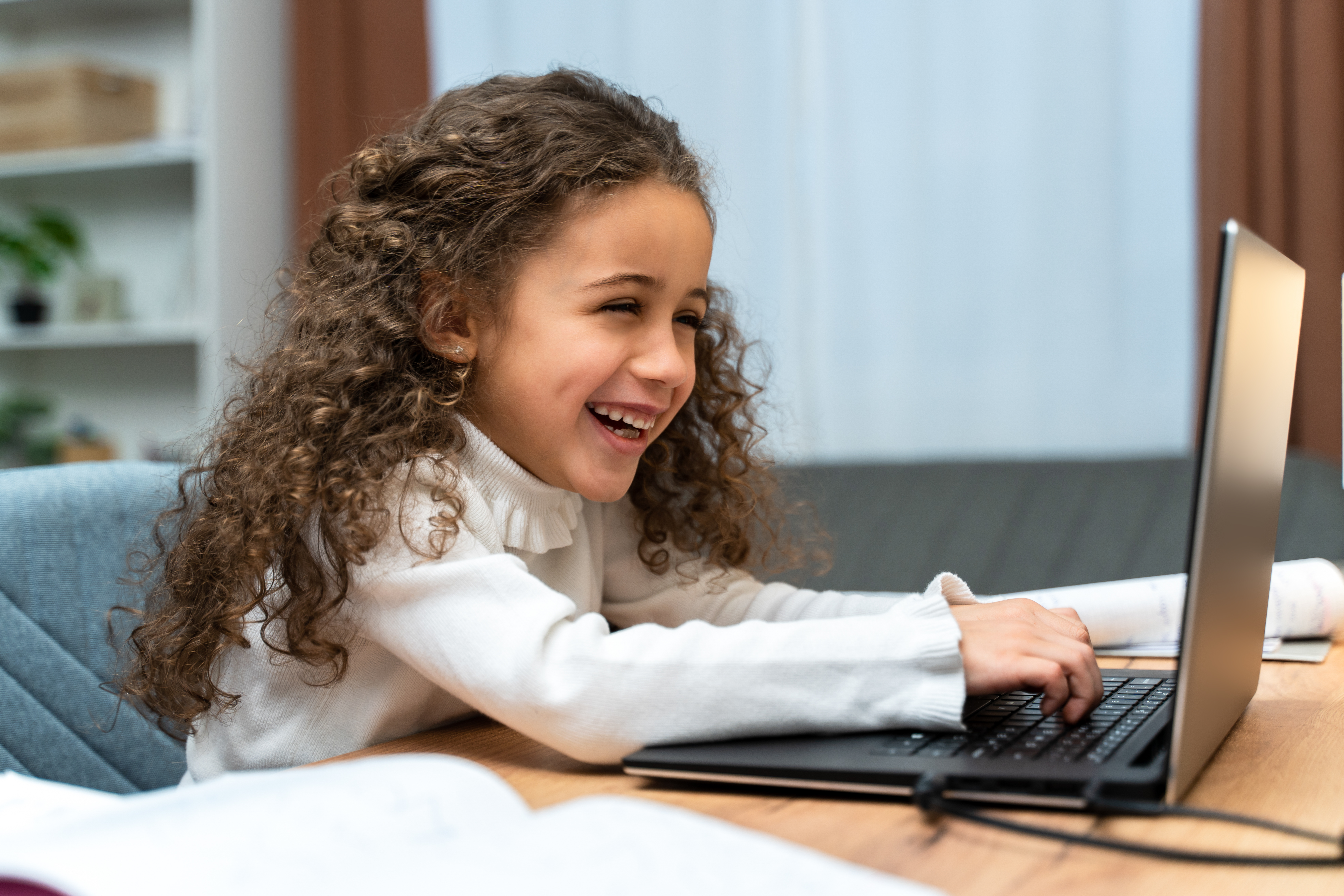 A young girl with long, curly brown hair is laughing joyfully while seated at a desk and typing on a black laptop, with an open book in the foreground.