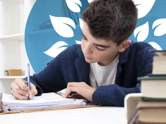 A homeschooling young boy with short black hair writing in a notepad at a desk with books.