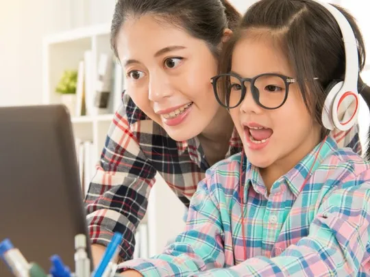 Smiling parent supporting child wearing headphones during an online lesson
