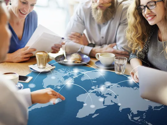 A diverse group of four adults sitting at a table with coffee, looking at an overlay graphic of a world map with connected glow lines.