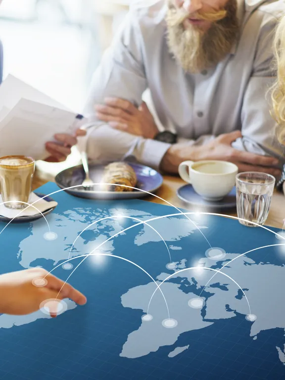 A diverse group of four adults sitting at a table with coffee, looking at an overlay graphic of a world map with connected glow lines.