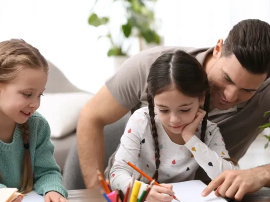 A dad homeschooling his two young daughters at a desk.