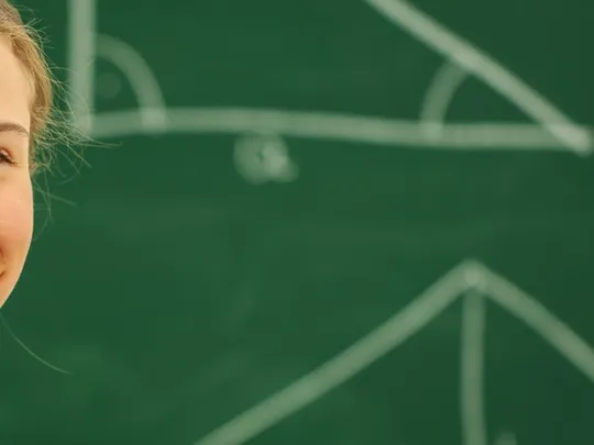 Confident girl standing in front of a green chalkboard with geometric shapes drawn behind her.