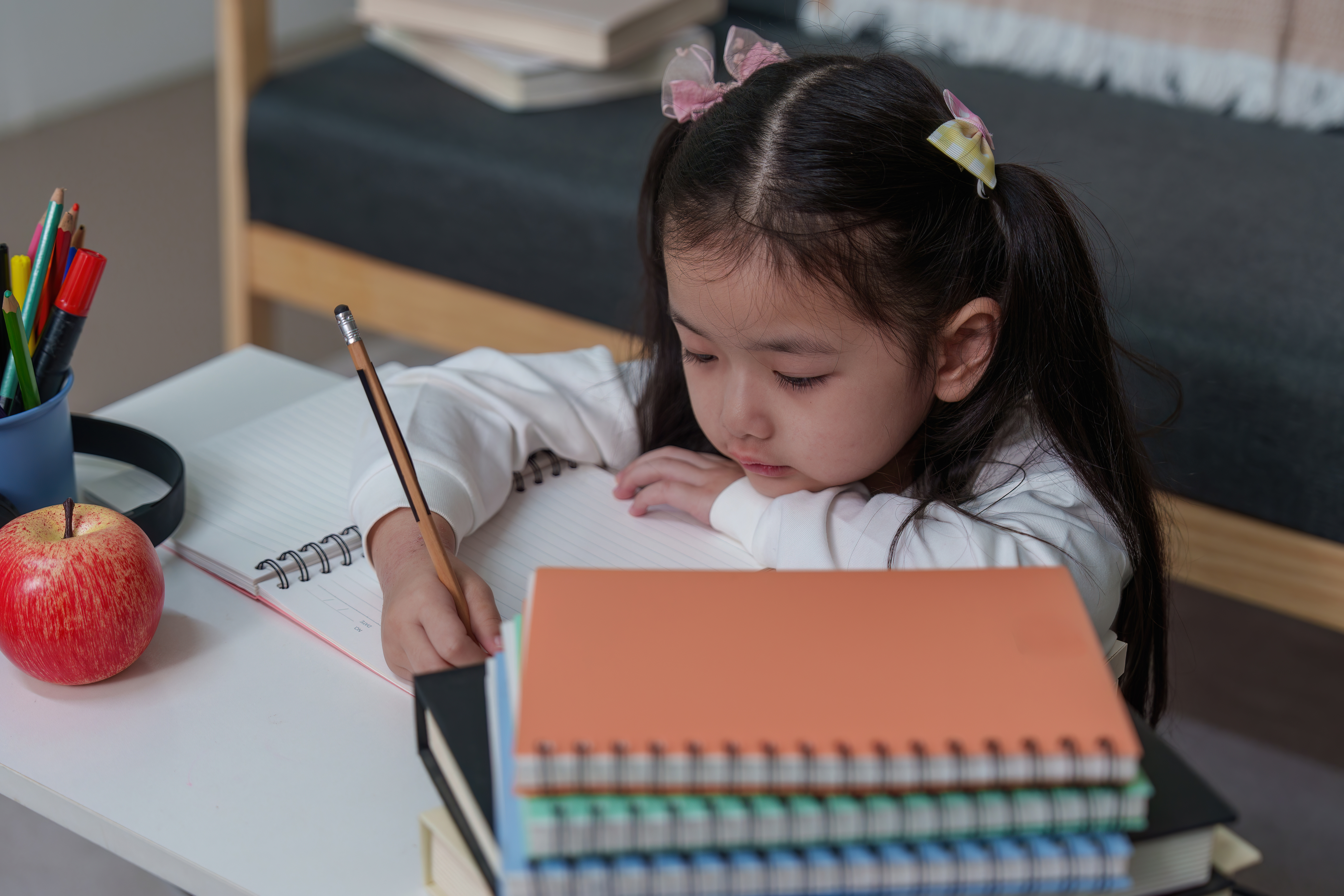 A young student with pigtails is focused on writing in a spiral notebook with a pencil, sitting at a desk next to a red apple and a stack of colorful books.