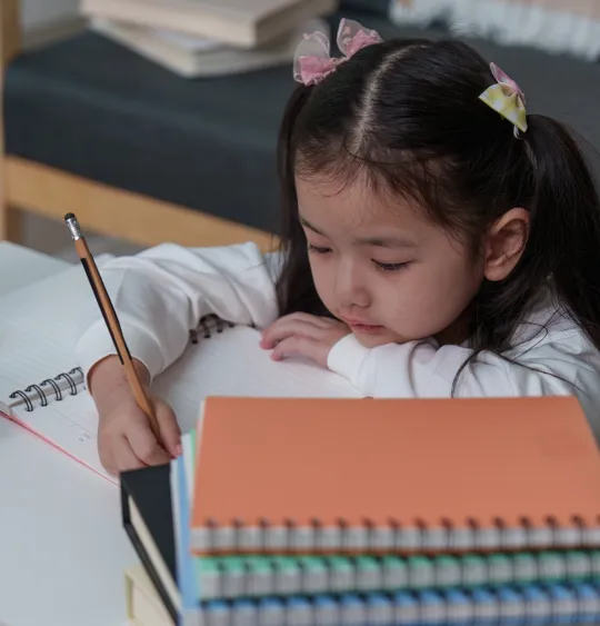 A young student with pigtails is focused on writing in a spiral notebook with a pencil, sitting at a desk next to a red apple and a stack of colorful books.