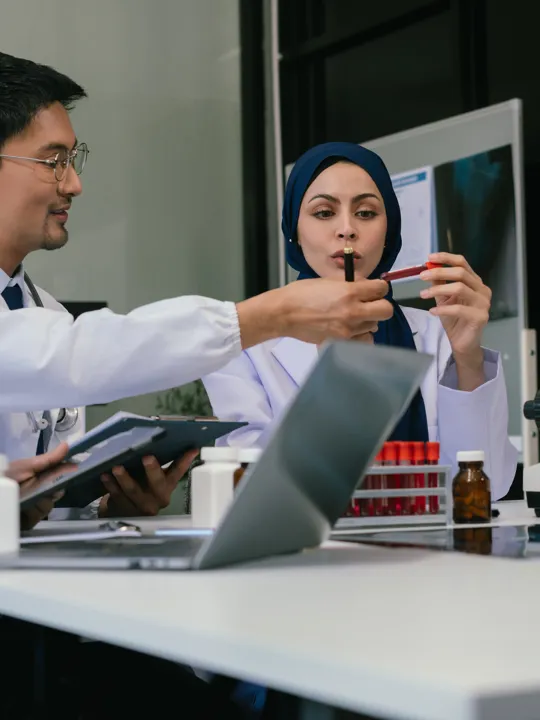 A team of four diverse scientists and medical professionals collaborating at a table with a microscope, test tubes, and a laptop, in a brightly lit laboratory setting.