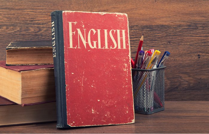 Worn red English textbook standing upright on a wooden desk next to a pencil holder and books.