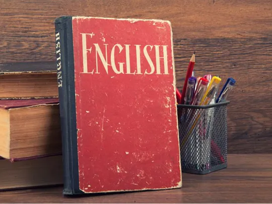 Worn red English textbook standing upright on a wooden desk next to a pencil holder and books.