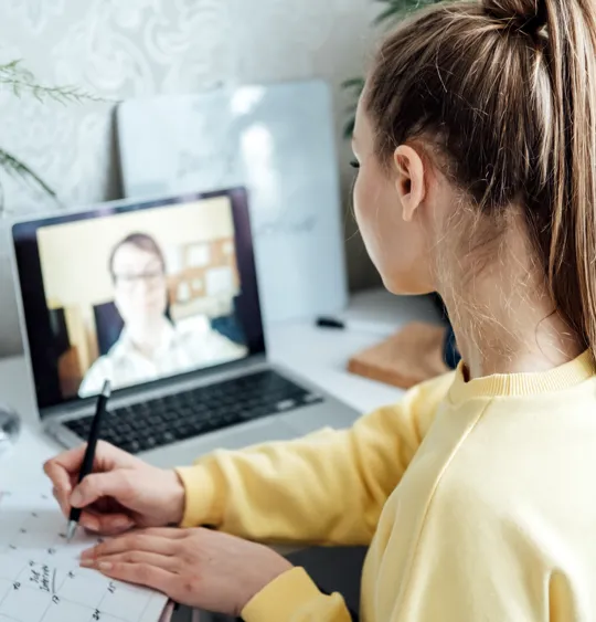 Student taking notes during a virtual lesson with a tutor on a laptop screen