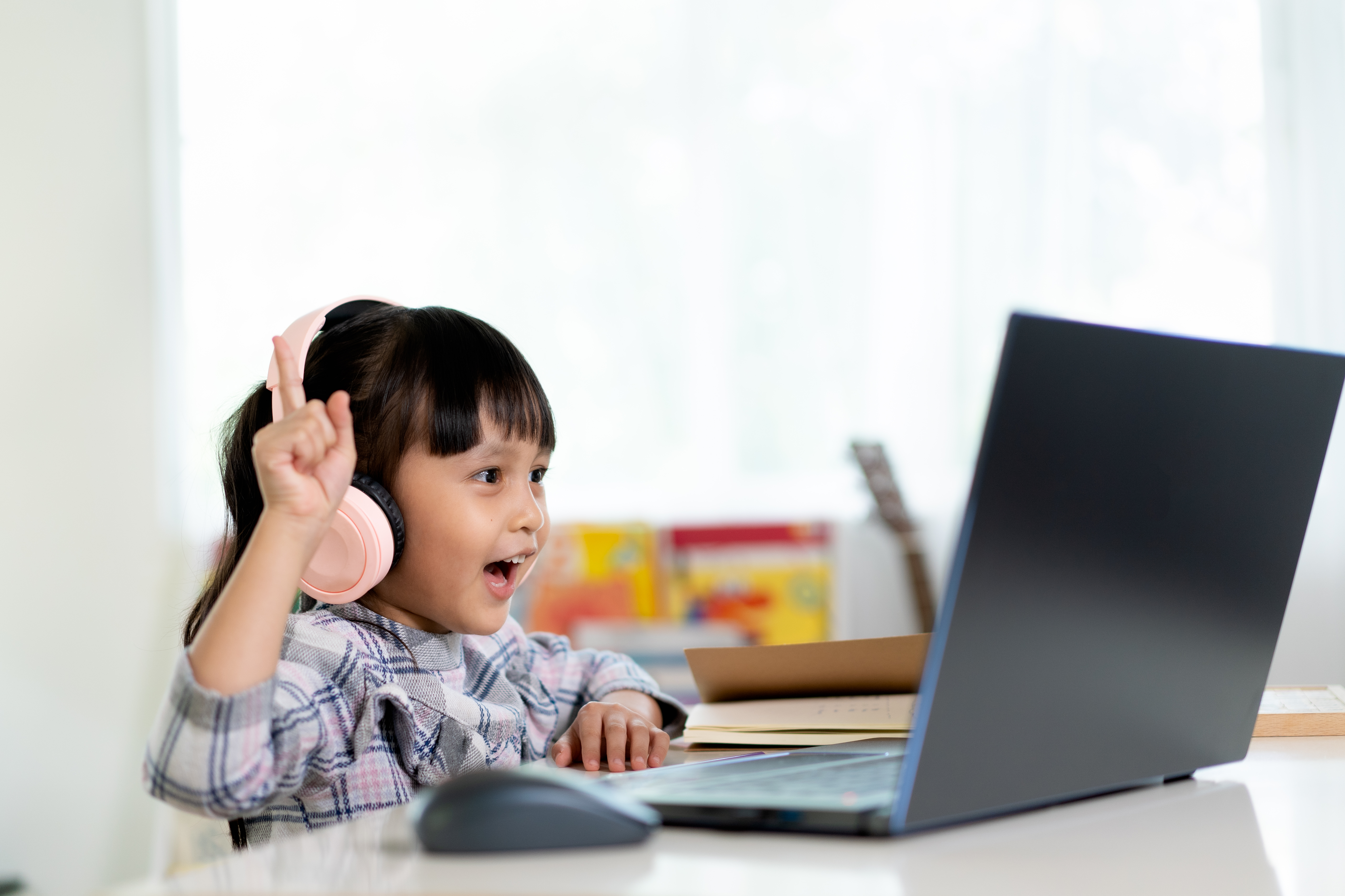 A young student is enthusiastically engaged in an online lesson, wearing light pink headphones and raising one hand while looking at a laptop screen.