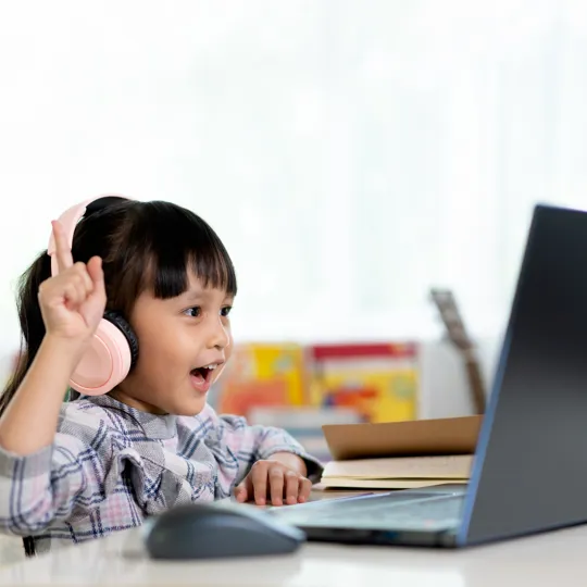 A young student is enthusiastically engaged in an online lesson, wearing light pink headphones and raising one hand while looking at a laptop screen.