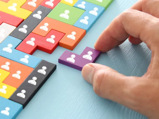 Hand placing a purple puzzle piece among colourful blocks, each marked with a white person icon
