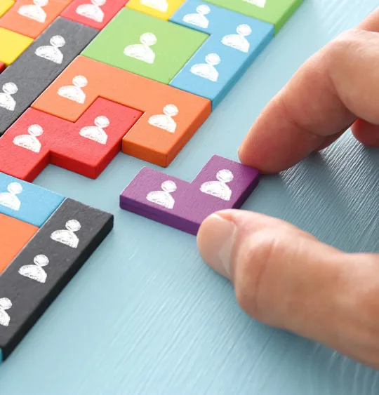Hand placing a purple puzzle piece among colourful blocks, each marked with a white person icon