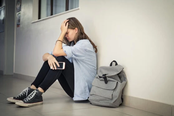 A young girl sitting against a wall with her head in her hand, and her phone in the other. She is looking anxious and upset.