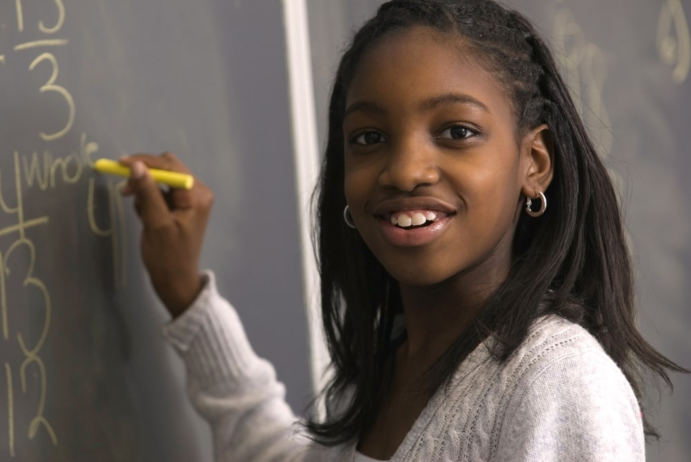 Schoolgirl solving a maths problem on a chalkboard, holding yellow chalk and smiling.