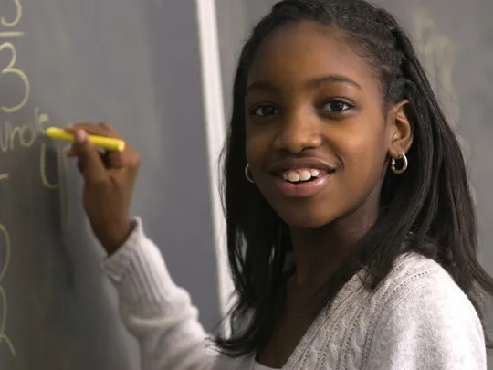 Schoolgirl solving a maths problem on a chalkboard, holding yellow chalk and smiling.